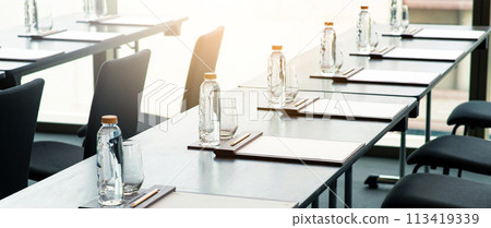 Empty seminar or business meeting room in the hotel. Plastic water bottles, Drinking glasses with pencils and paper notes set up on the table prepared for conferences or meetings. Empty seminar or business meeting room in the hotel. Plastic water bottles, Drinking glasses with pencils and paper notes set up on the table prepared for conferences or meetings. 113419339