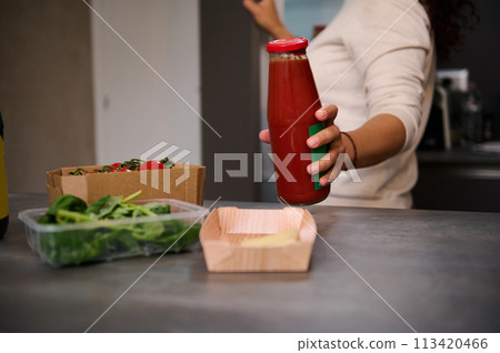 Close-up woman putting bottle of tomato juice on kitchen counter with fresh ingredients for salad. Housewife taking out ingredients from the fridge, preparing healthy meal at home kitchen Close-up woman putting bottle of tomato juice on kitchen counter with fresh ingredients for salad. Housewife taking out ingredients from the fridge, preparing healthy meal at home kitchen 113420466