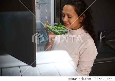 Charming multi ethnic woman holding box with fresh arugula leaves, smelling it, standing by open fridge in the home kitchen. Charming multi ethnic woman holding box with fresh arugula leaves, smelling it, standing by open fridge in the home kitchen. 113420468