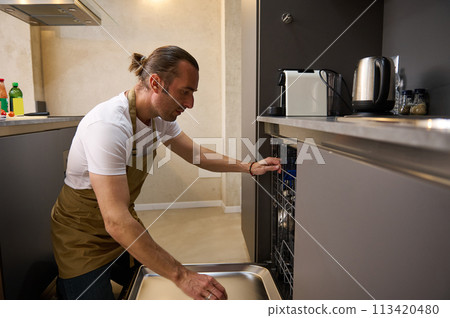 Handsome man in beige chef's apron, emptying out, loading or unloading, taking clean dishes out of a modern automatic dishwasher at minimalist home kitchen interior. People. Household chores Handsome man in beige chef's apron, emptying out, loading or unloading, taking clean dishes out of a modern automatic dishwasher at minimalist home kitchen interior. People. Household chores 113420480