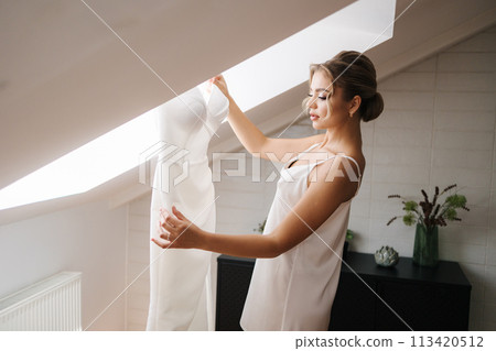 A beautiful bride in a silk white robe stands by her wedding dress hanging on the skylight A beautiful bride in a silk white robe stands by her wedding dress hanging on the skylight 113420512