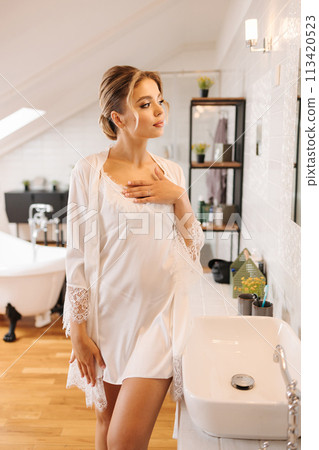 Beautiful and tender bride in white silk robe standing in luxury bathroom. Female preparing for wedding Beautiful and tender bride in white silk robe standing in luxury bathroom. Female preparing for wedding 113420523