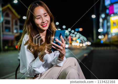 Asian woman on a night walk through the city streets multitasks with technology by using a laptop computer and mobile phone while talking. 113422168