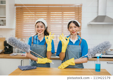 Two Asian young cleaning service women on kitchen counter with duster foggy spray and rag showcase efficient housework teamwork. Clean portrait two uniform maid working smiling employee. 113422179