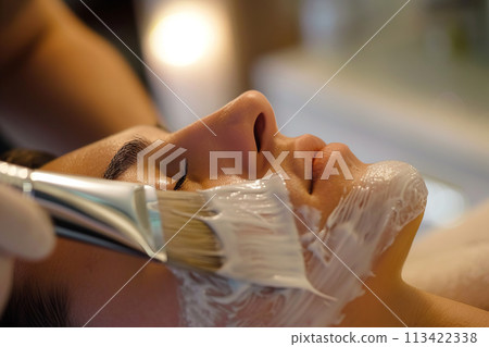 A young girl undergoes a spa procedure of applying a mask to her face from a cosmetologist and relaxes 113422338