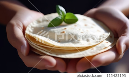 Hands delicately holding a stack of warm, soft tortillas adorned with a basil leaf. 113423193