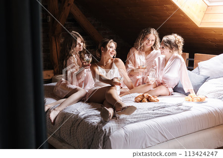 Four girl sitting on the bed and drinking champagne from glasses. Bridesmaids smile and have fun before wedding. Croissants and other snacks 113424247