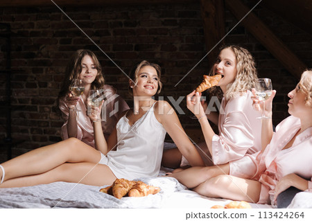 Portrait of four females sitting on bed and eating croissants. Bridesmaids and bride wears silk white robe. Woman drinks champagne 113424264