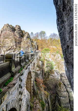 A senior traveler with a backpack and trekking poles walks on the Bastei Bridge amid sandstone formations in Saxon Switzerland National Park. Germany 113426692