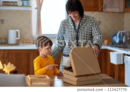 Grandmother and grandson are standing at the table and opening a box of pizza. Generation connection concept. Grandmother and grandson are standing at the table and opening a box of pizza. Generation connection concept. 113427776