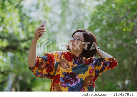 Cheerful young lady with glasses capturing a selfie in a lush green park. Young entrepreneur with digital devices capturing a selfie, staying connected in an urban green space Cheerful young lady with glasses capturing a selfie in a lush green park. Young entrepreneur with digital devices capturing a selfie, staying connected in an urban green space 113427951