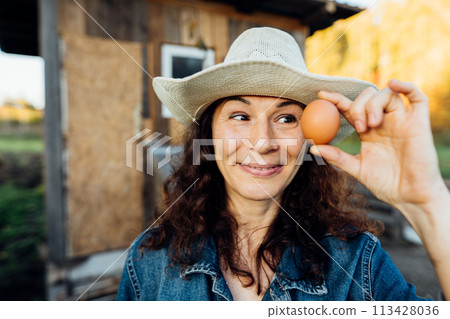 Portrait of funny joyful farmer woman holding fresh egg near her face. Portrait of a funny farmer with an egg in the summer. Happy moment of rural life Portrait of funny joyful farmer woman holding fresh egg near her face. Portrait of a funny farmer with an egg in the summer. Happy moment of rural life 113428036