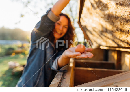 Country joy, woman with an egg in a country chicken coop. Cheerful homesteader showcases an egg at her countryside dwelling. Sustainable lifestyle, organic farmer 113428046