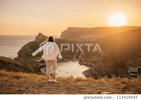 A woman stands on a hill overlooking a body of water. The sun is setting in the background, casting a warm glow over the scene. The woman is enjoying the view and taking in the beauty of the landscape A woman stands on a hill overlooking a body of water. The sun is setting in the background, casting a warm glow over the scene. The woman is enjoying the view and taking in the beauty of the landscape 113429597