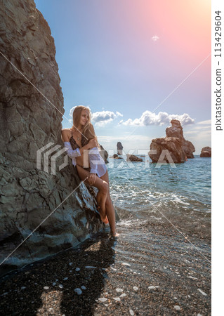 Woman travel sea. Attractive blonde woman in a black swimsuit enjoying the sea air on the seashore around the rocks. Travel and vacation concept. 113429604