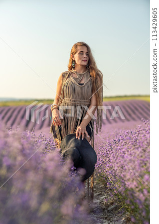 A woman is standing in a field of purple flowers, wearing a black dress and a black hat. 113429605