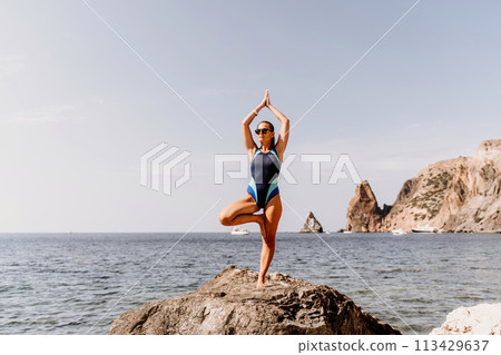 Yoga on the beach. A happy woman meditating in a yoga pose on the beach, surrounded by the ocean and rock mountains, promoting a healthy lifestyle outdoors in nature, and inspiring fitness concept. Yoga on the beach. A happy woman meditating in a yoga pose on the beach, surrounded by the ocean and rock mountains, promoting a healthy lifestyle outdoors in nature, and inspiring fitness concept. 113429637
