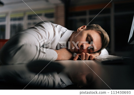 Closeup of tired sleepy male employee asleep on computer desk late at night. Exhausted businessman sleeping with head on keyboard while working overnight on computer. Concept overwork and deadline. Closeup of tired sleepy male employee asleep on computer desk late at night. Exhausted businessman sleeping with head on keyboard while working overnight on computer. Concept overwork and deadline. 113430693