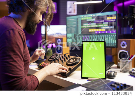 Composer creating soundtracks on stereo gear and greenscreen display on mobile device, mixing and mastering tunes in home studio. Young artist adjusting volume levels on music, daw software. 113433934