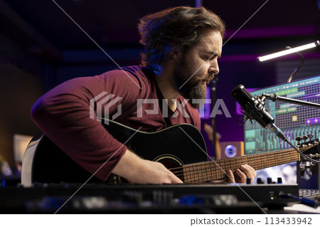 Songwriter composing a new song on acoustic guitar in home studio, using a microphone to sing the chords. Artist works with mixing console and daw software, guitarist producing new music. 113433942