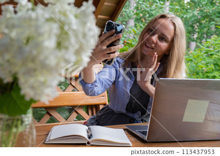 Young happy woman making video call on smartphone in wooden alcove. Relaxed outdoor setting emphasizes comfort and productivity. Remote work learning 113437953