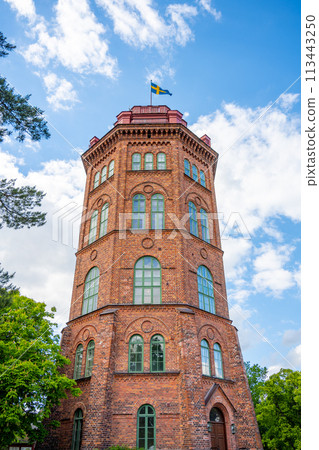 Bredablick Tower stands tall against a blue, cloud-speckled sky at Skansen in Stockholm, Sweden, showcasing its brick architecture and Swedish flag. Bredablick Tower stands tall against a blue, cloud-speckled sky at Skansen in Stockholm, Sweden, showcasing its brick architecture and Swedish flag. 113443250