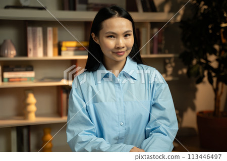 Smiling Young Woman in Blue Shirt Standing in a Cozy Home Library 113446497