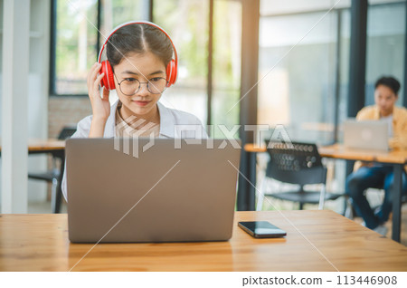 Student using a laptop in a cafe while wearing headphones, learning and searching the internet, and making a video call. 113446908