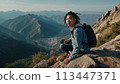 A young happy Chinese man with a tourist backpack stands on top of a rock in the mountains, travels and enjoys a beautiful view of the mountains, sky or nature. Untouched nature. Tourism and travel 113447371