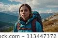 A young happy European woman with a tourist backpack stands on top of a rock in the mountains, travels and enjoys a beautiful view of the mountains, sky or nature. Untouched nature. Tourism and travel 113447372