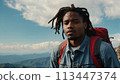 Young positive African American man with a tourist backpack stands on top of a rock in the mountains, travels and enjoys a beautiful view of the mountains, sky or nature. Untouched nature. Tourism and 113447374
