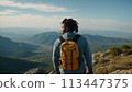 Young positive African American man with a tourist backpack stands on top of a rock in the mountains, travels and enjoys a beautiful view of the mountains, sky or nature. Untouched nature. Tourism and 113447375