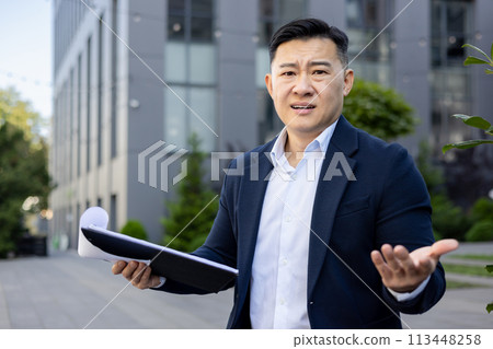 Close-up portrait of a young Asian man in a business suit standing outside an office building, holding documents and looking worriedly at the camera. Close-up portrait of a young Asian man in a business suit standing outside an office building, holding documents and looking worriedly at the camera. 113448258