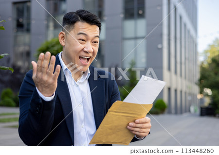 Happy and surprised young asian male businessman standing outside on the street in a hat and celebrating reading a received letter in an envelope. 113448260