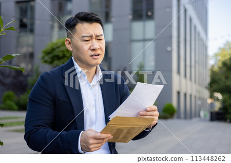 Upset and disappointed Asian businessman sitting outside the office and reading a received letter with bad news. Upset and disappointed Asian businessman sitting outside the office and reading a received letter with bad news. 113448262