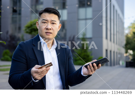 Close-up portrait of young worried Asian man businessman sitting near office building, holding phone and credit card in hands and looking upset at camera, financial problems with account. Close-up portrait of young worried Asian man businessman sitting near office building, holding phone and credit card in hands and looking upset at camera, financial problems with account. 113448269
