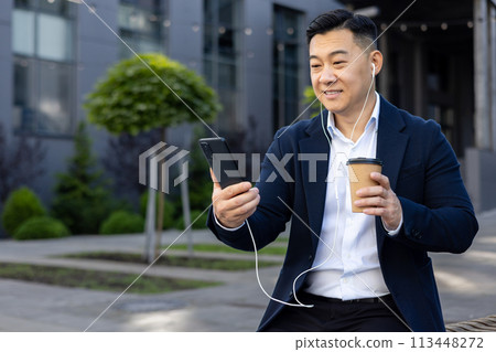 Asian young male office worker sitting outside an office building wearing headphones, talking on the phone on a video call and drinking coffee. Asian young male office worker sitting outside an office building wearing headphones, talking on the phone on a video call and drinking coffee. 113448272