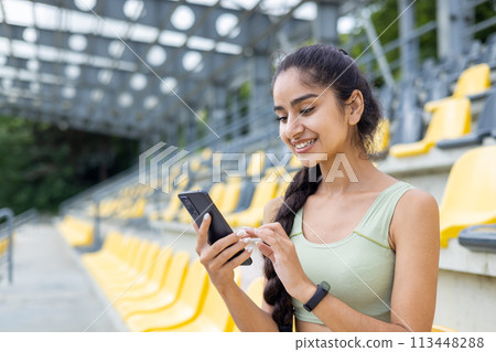 Close-up photo of a smiling young Indian girl who is in a stadium and using a mobile phone. 113448288
