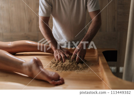 Cropped shot of unrecognizable massage practitioner male mixing heated sand on massage table for scrubbing treatment in luxury spa salon. Preparation to serenity of recreation in wellness center. 113449382