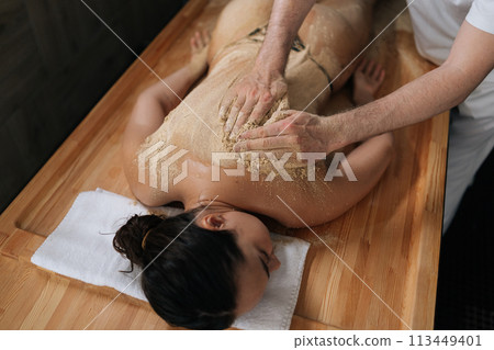 High-angle view of unrecognizable masseur male massaging with scrub of back for female client in wellness center. Serene young woman lying enjoying back scrub at luxury spa salon with cozy light. 113449401