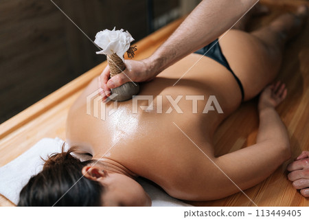High-angle view of relaxed young woman lying with closed eyes receiving herbal bolus bags massage. Closeup hands of Ayurveda massage therapist pressing herbal bolus bags onto female body. 113449405