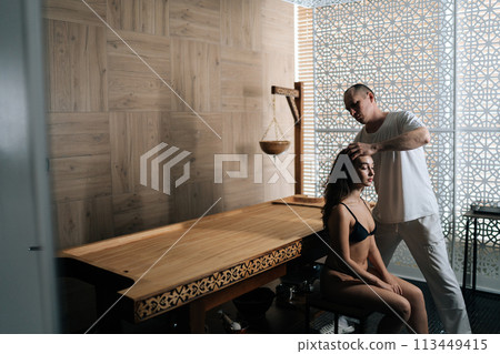 Remote view of masseur male make relaxing therapeutic head massage to young woman wearing underwear in wellness center. Happy pretty female getting head massage by professional masseur in spa salon. Remote view of masseur male make relaxing therapeutic head massage to young woman wearing underwear in wellness center. Happy pretty female getting head massage by professional masseur in spa salon. 113449415