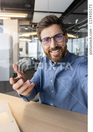Happy bearded man in a blue shirt reaching towards the camera, offering a greeting with a warm, friendly smile in an office setting. 113450173