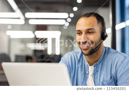 Close-up photo of a smiling young Hispanic man sitting in the office in front of a laptop monitor and working in a headset. 113450201