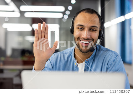 Close-up photo of a young smiling Arab man in a headset talking on a video call on a laptop while waving his, hand at the camera. 113450207