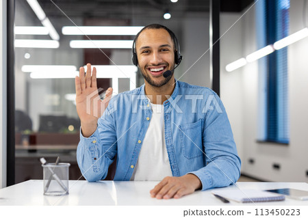Smiling young African American man in headset sitting in office in front of camera, talking and waving hand to camera. 113450223