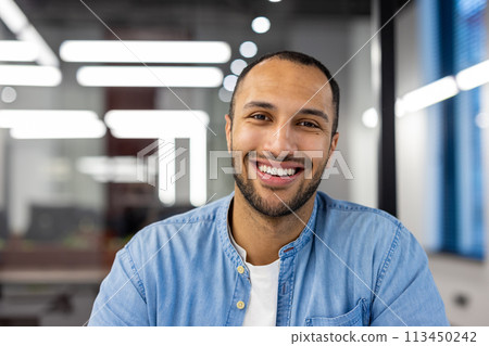 Focused Indian man at home in his living room, engaged in a business call, looking professionally into the camera in a casual setting. Focused Indian man at home in his living room, engaged in a business call, looking professionally into the camera in a casual setting. 113450242