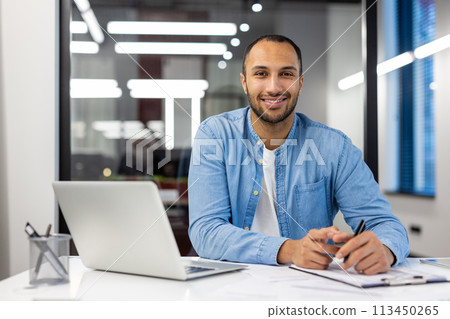 Confident Indian man with a friendly smile working on a laptop in a well-lit contemporary office setting. Confident Indian man with a friendly smile working on a laptop in a well-lit contemporary office setting. 113450265