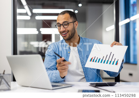 Smiling Indian businessman in casual wear holding a bar chart report during a presentation in a modern office setting. Smiling Indian businessman in casual wear holding a bar chart report during a presentation in a modern office setting. 113450298