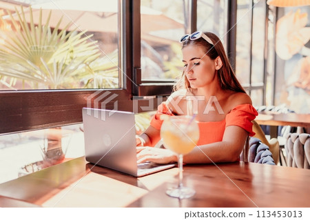 Woman works on laptop in a sunny cafe, glass of juice nearby. The setting is modern, with natural light illuminating the space. The image captures a moment of focused work. Woman works on laptop in a sunny cafe, glass of juice nearby. The setting is modern, with natural light illuminating the space. The image captures a moment of focused work. 113453013